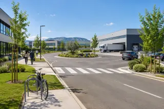 Modern business park entrance with light industrial buildings, a truck at a loading dock, office workers on a sunny day, clean roads and landscaped greenery.
